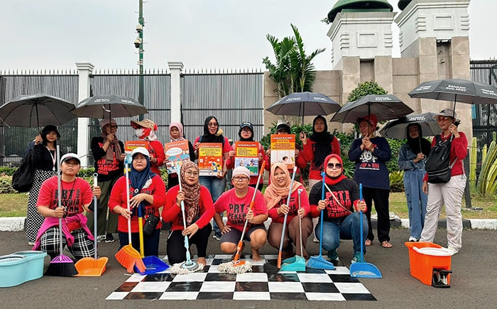 Die vordere Reihe einer Gruppe von Hausangestellten kniet und hält Putzgeräte, die dahinter Stehenden halten Plakate und beschriftete Regenschirme. Falls alle tragen rote T-Shirts.