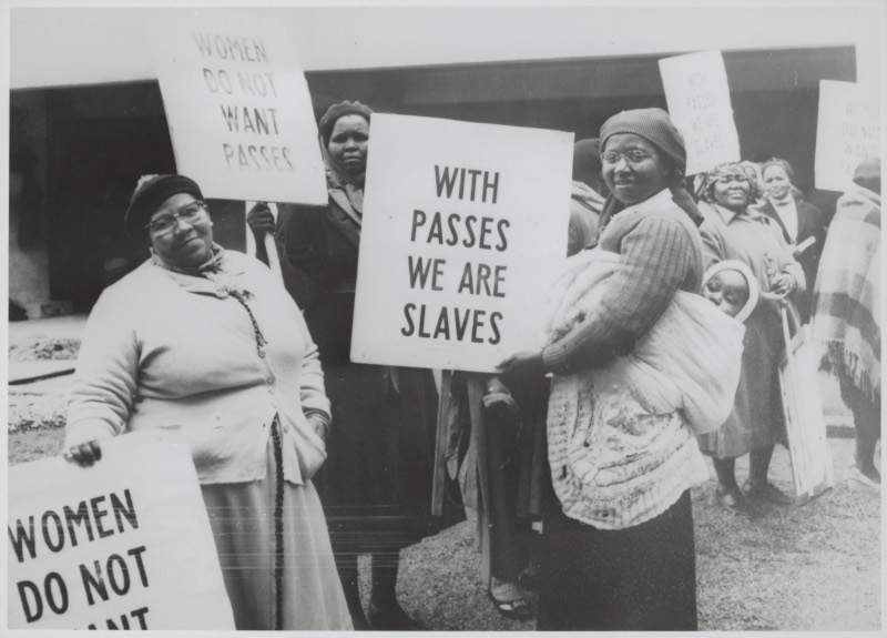 Mehrere Schwarze Frauen, davon eine mit einem Kleinkind in einem Tuch auf dem Rücken, halten Schilder mit Texten: „Women do not want passes“ oder „With passes we are slaves“.